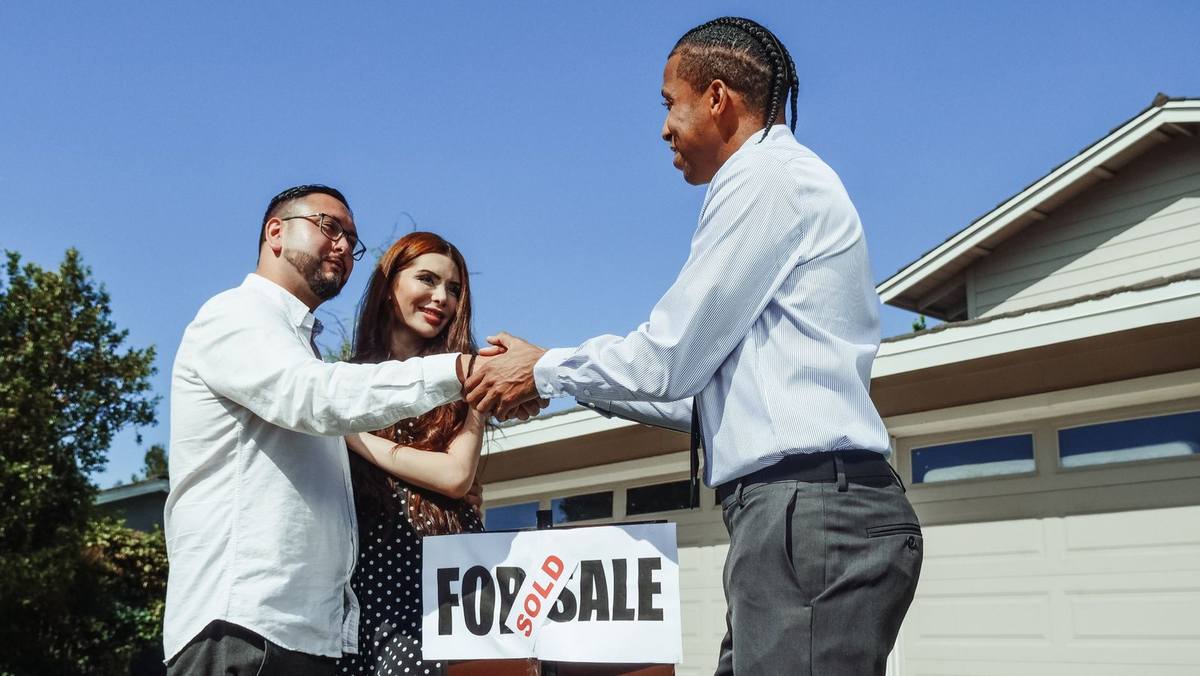 Homme saluant un couple devant une maison avec panneau 