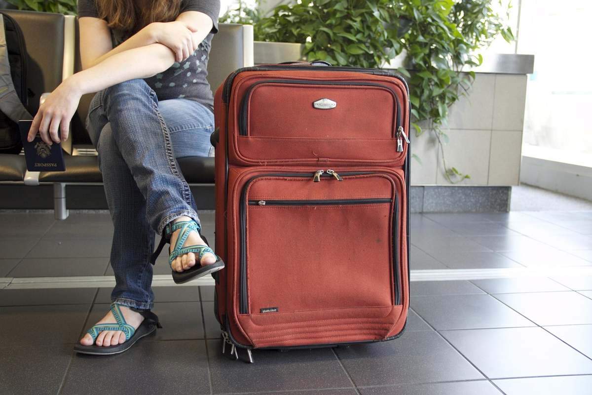 Femme en jeans et sandales, valise rouge à roulettes, siège public, végétation derrière